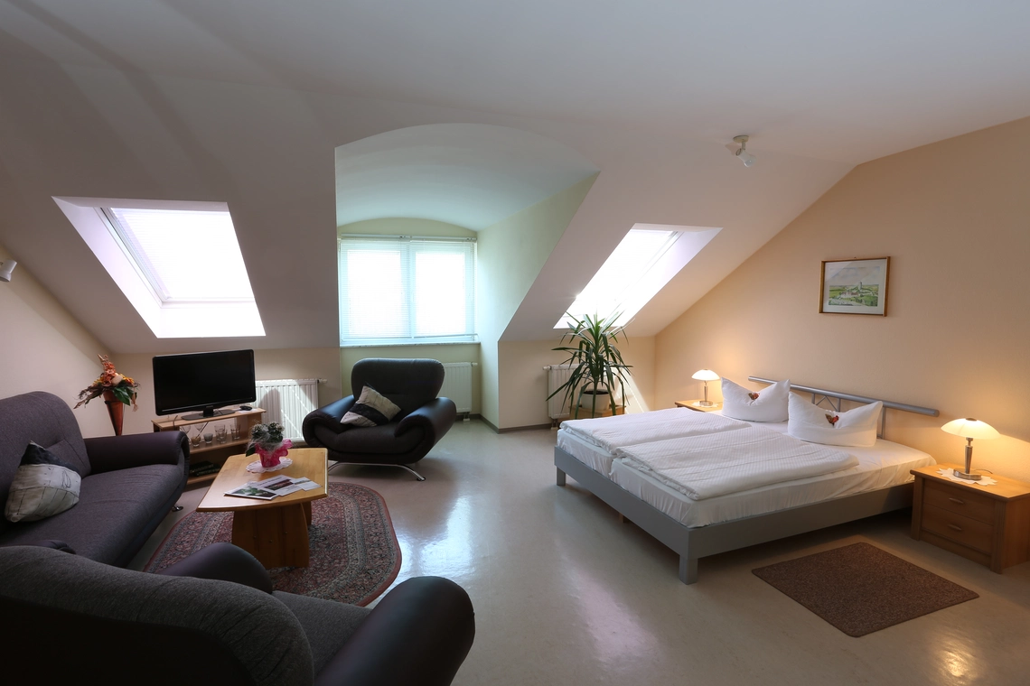 Ein Gästezimmer mit Doppelbett und schwarzer Couch und Sesseln, Breitbildfernseher und Blumendekoration.