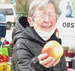 Eine Bewohnerin ist auf dem Wochenmarkt und hält einen Apfel in die Kamera.