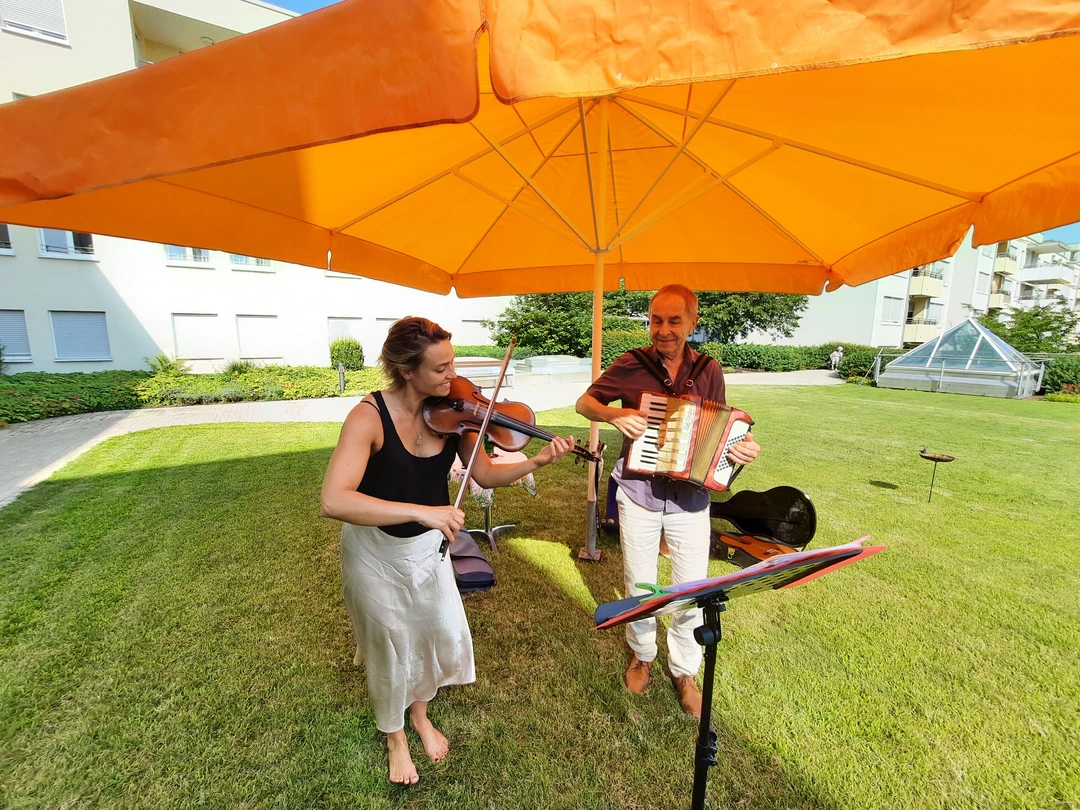 Musiker mit Akkordeon und Musikerin mit Geige unter einem Sonnenschirm in Orange.