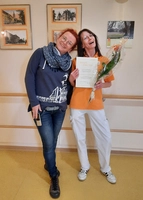 Heimleiterin Daniela Müller überreicht Pflegehelferin Nicole Planitzer eine Urkunde und Blumen, beide lehnen sich aneinander und lachen.