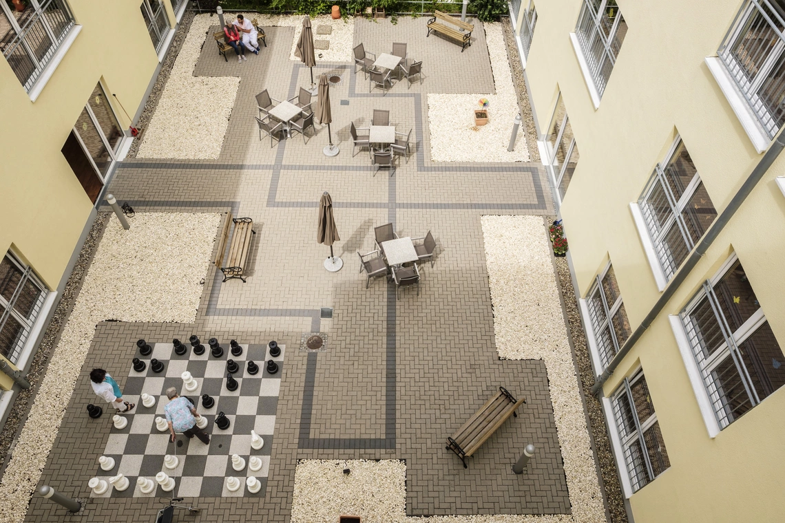 Fotografie des Innenhofs vom 3. OG aus; man sieht ein großes Schachspiel mit tragbaren Figuren, außerdem Tische, Stühle, Bänke und Sonnenschirme.