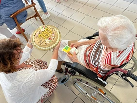 Eine Mitarbeiterin kniet neben dem Rollstuhl einer Bewohnerin und überreicht ihr zum 100. Geburtstag eine Torte.