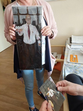 Eine Mitarbeiterin hält das von Ali gemalte Porträt eines Bewohners in der Hand. Es zeigt ihn beim Kellnern. Auch die Foto-Vorlage ist zu sehen.