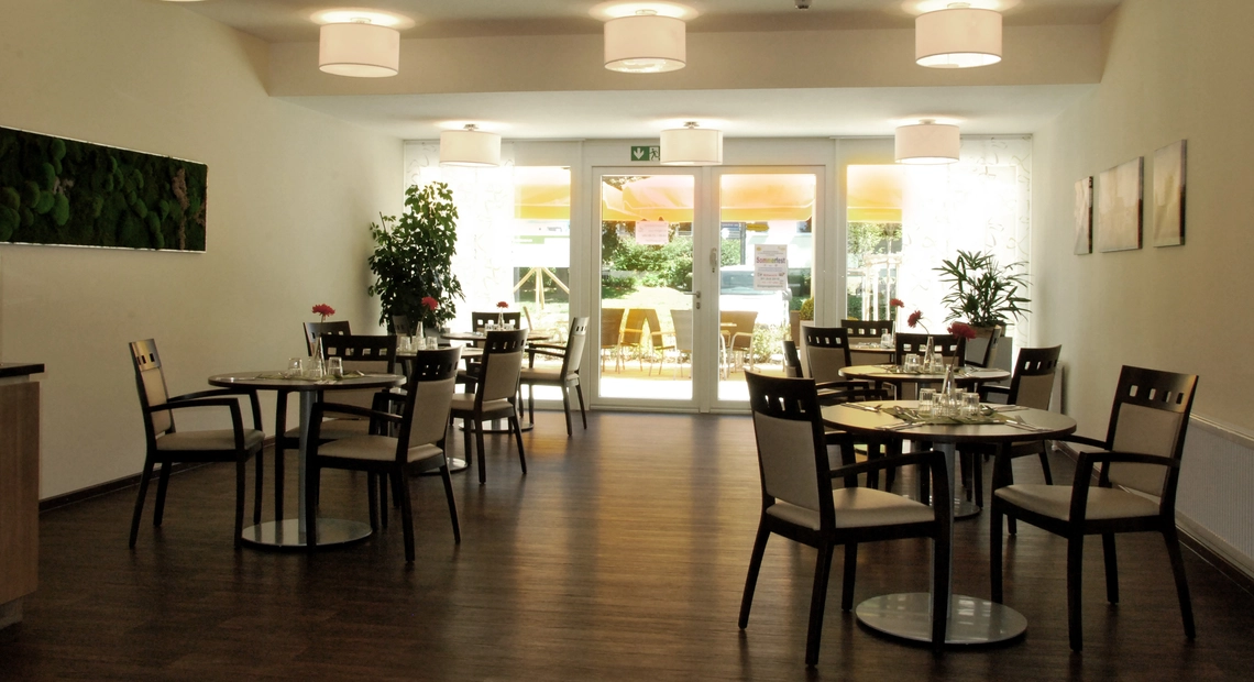 Caféteria Innenraum mit braunen Tischen und Stühlen auf braunem Holzboden, Blick durch große Fenster nach außen auf die Terrasse.