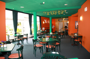 Das leere Café, links große Panoramafenster, Decke und Möbel grün, Wände orange, auf den Tischen stehen kleine Blumenvasen.