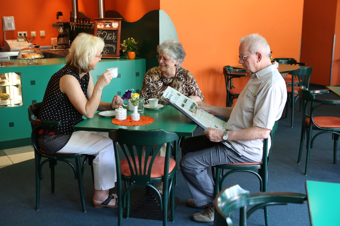 Café mit grünen Tischen und Stühlen, ein älterer Herr liest Zeitung, zwei Frauen am selben Tisch unterhalten sich amüsiert beim Kaffeetrinken.