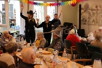 Musiker im Hintergrund, Mitarbeiterin tanzt mit Bewohnerin, Publikum an Tischen in der Cafeteria