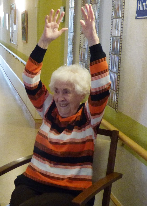Eine Senioren sitzt auf einem Stuhl und streckt die Hände in die Höhe.