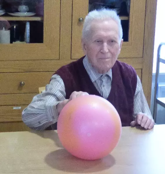 Ein alter Mann sitzt am Tisch und rollt einen roten Ball mit der linken Hand weiter zu seinem Gegenüber.