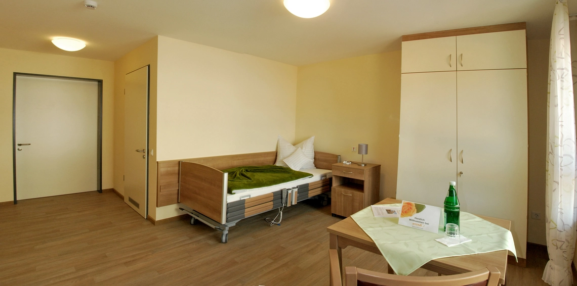 Pflegezimmer mit elektrisch verstellbarem Bett, dunkelbraunem Parkett, weißem Schrank, einem Holztisch und einem Stuhl.