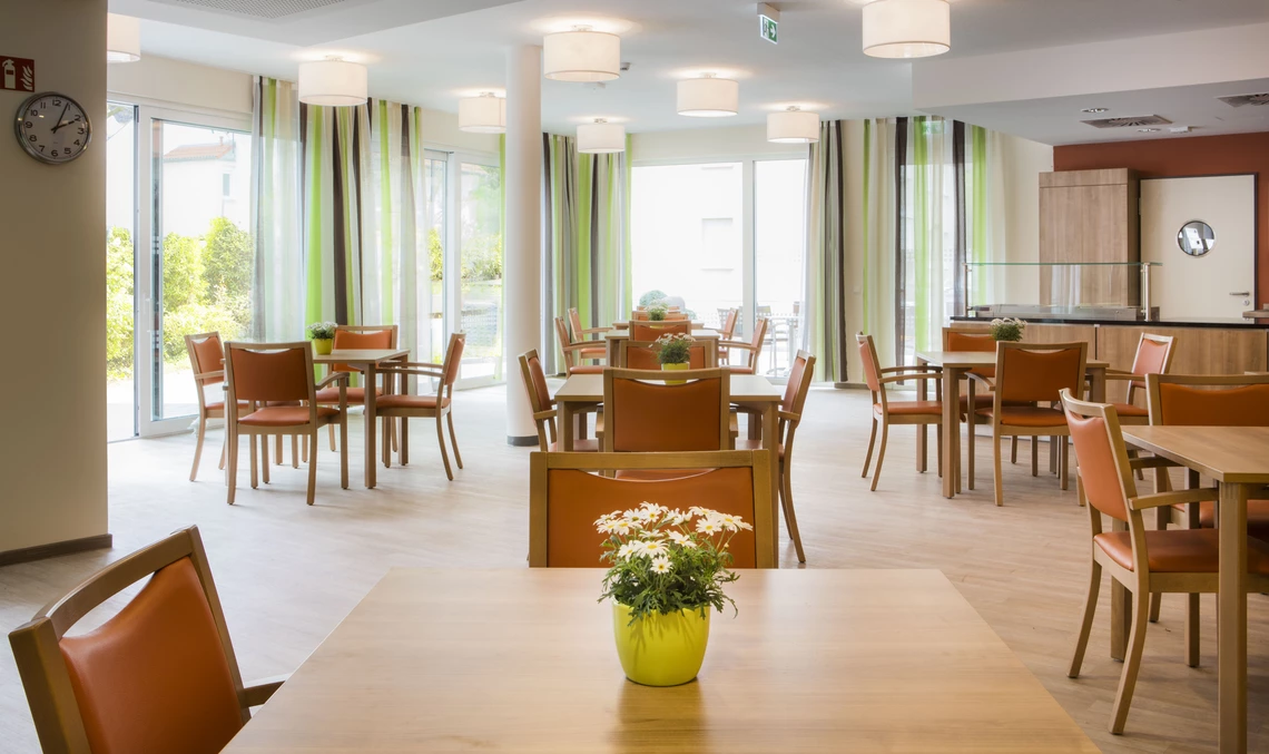 Cafeteria mit mehreren Holztischen, pro Tisch eine Blumenvase, große Fenster mit Vorhängen.