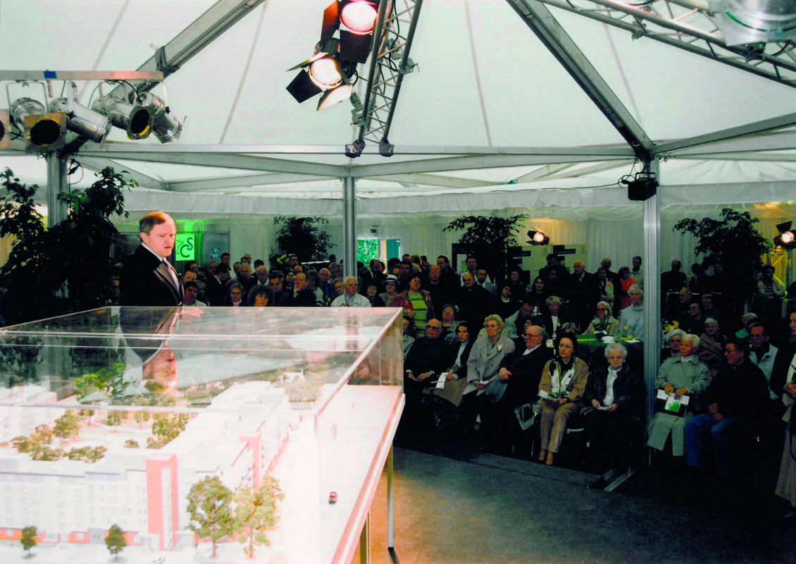 Rede von Heinz Scheidel bei der Premiere des Lanzgartens. Er blickt auf einen Glaskasten mit einem Modell der Einrichtung. Das Publikum sitzt bzw. steht und hört zu.