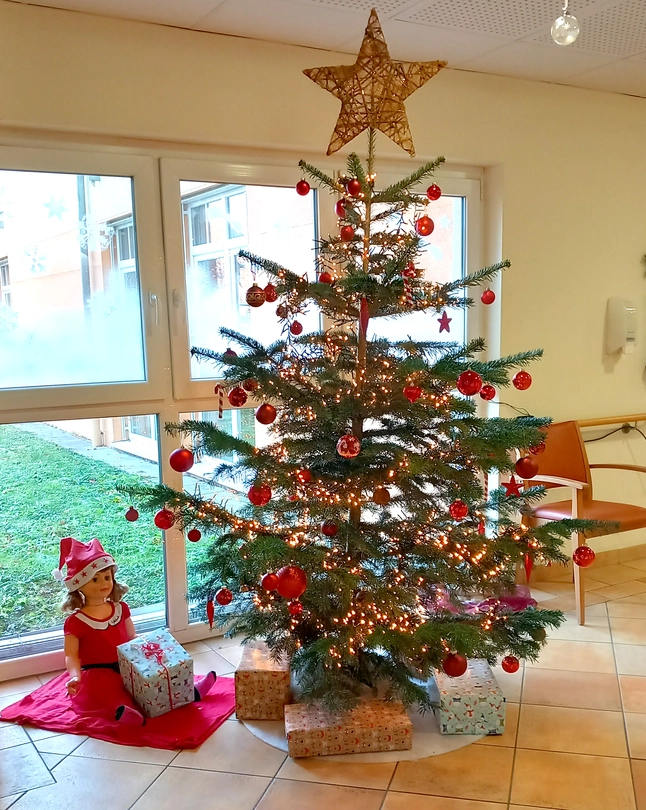 Ein geschmückter Christbaum mit Geschenken darunter, links sitzt eine große Puppe mit einem Geschenk im Schoß