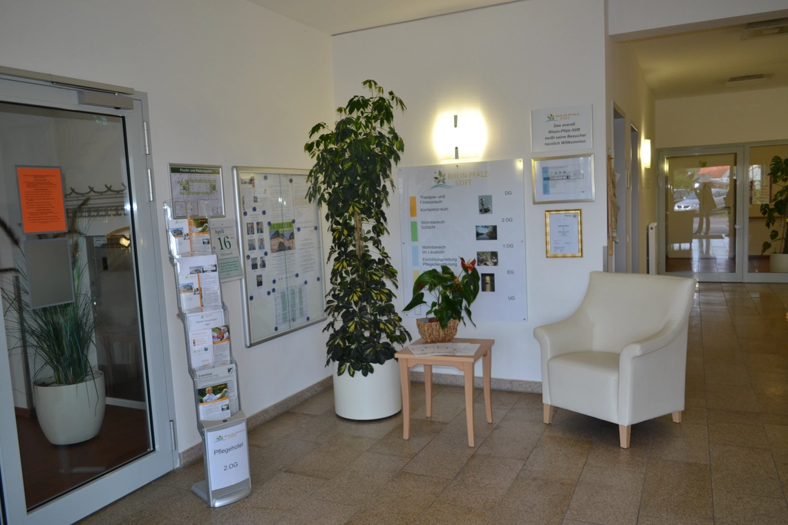 Rhein-Pfalz-Stift Eingangshalle mit Holztisch und weißem Sessel, Pinwand, Wegweiser, Prospektständer und Topfpflanze.