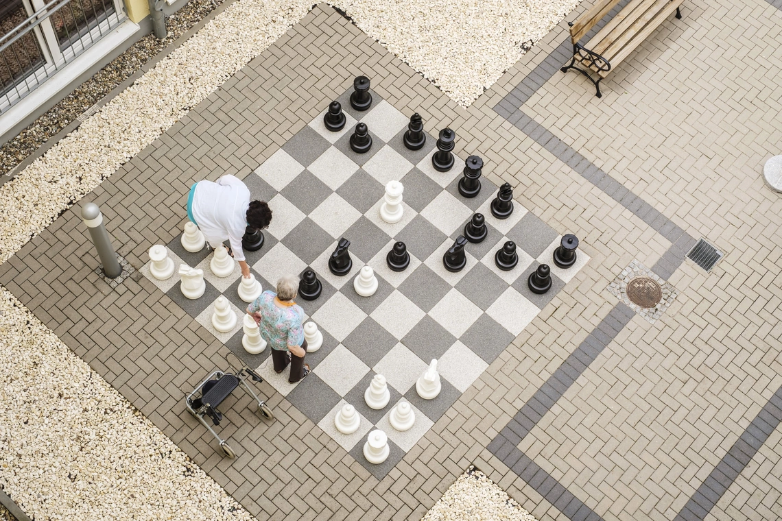 Eine Bewohnerin und eine Pflegerin spielen im Innenhof Schach auf einem Schachfeld aus Steinplatten