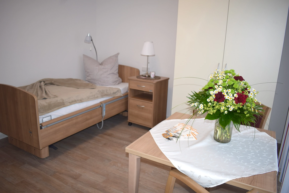 Wohnraum mit hölzernem Bett und Nachttisch, einem Schrank und einem Tisch mit Decke und Blumenvase darauf