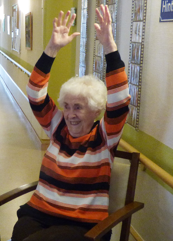 Eine Senioren sitzt auf einem Stuhl und streckt die Hände in die Höhe.