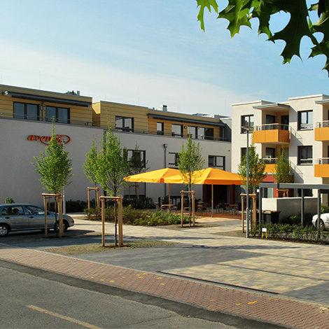 Standort An der Wiesenau, großer Parkplatz, Blick auf Terrasse mit orangenen großen Sonnenschirmen, orangefarbene Balkone.
