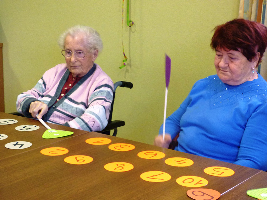 Zwei Seniorinnen halten Fliegenklatschen in der Hand, auf dem Tisch vor ihnen liegen Zahlen-Karten.